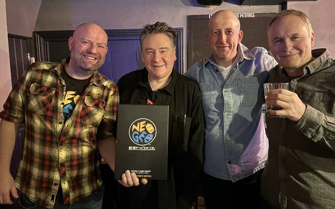 Four smiling white men gathered in a pub. One is holding a pint of beer. Another is holding a large black book.